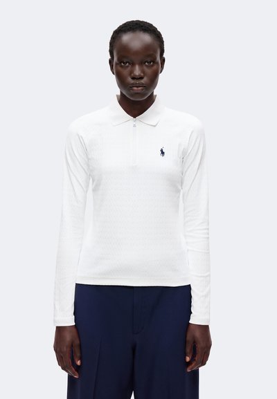 Femme portant un polo blanc à manches longues avec un logo bleu marine et un pantalon bleu marine, debout devant un fond clair uni.
