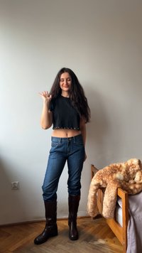 Czarny krótki top z ozdobnymi oczkami, niebieskie dżinsy oraz brązowe kozaki za kolano, stojąca obok łóżka z miękkim brązowym narzutą.