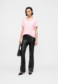 Γυναίκα που φοράει ροζ T-shirt με V λαιμόκοψη, μαύρο δερμάτινο παντελόνι, μαύρα μυτερά τακούνια, κρατώντας μια μαύρη τσάντα με τρουκ, στέκεται σε λευκό φόντο.