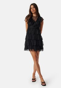 Robe en dentelle noire avec un design à volants, encolure transparente, accents froncés et manches courtes. Tissu léger avec une finition texturée.