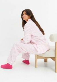 Set di pigiama a righe rosa e bianche con maniche lunghe e pantaloni, abbinato a calzini rosa pelosi. Seduto su una sedia in legno chiaro.