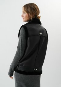 Czarna skórzana kamizelka o teksturowym wykończeniu, z futrzanym kołnierzem i srebrnym elementem ozdobnym z tyłu, połączona z szarym swetrem dzianym.