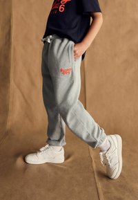Sivé bavlnené tepláky s oranžovým logom "Super Dry", elastickým pásom a rebrovanými manžetami, skombinované s bielymi teniskami na textúrovanom hnedom pozadí.