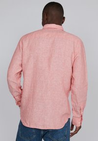 Svetlo roza lanena srajca z dolgimi rokavi, vidnimi šivi in gumbastim ovratnikom, oblečena z modrimi jeans hlačami.
