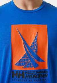 Camiseta de algodón azul con un gráfico en forma de cuadrado naranja que presenta un diseño de vela estilizada y texto en negro "HELLY HANSEN HYDRO PWR" en la parte inferior.