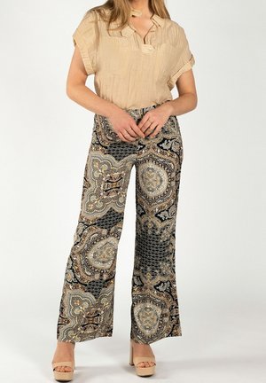 Mujer con blusa beige de manga corta metida en pantalones anchos con estampado, combinados con sandalias beige de tacón alto, de pie sobre fondo blanco.