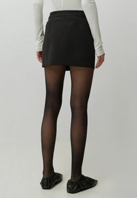 Zwarte mini rok met een gladde textuur en tailleband, gedragen met sheer zwarte panty’s en zwarte platte schoenen met een gerimpeld ontwerp.