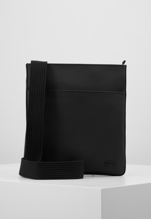 Sort tekstureret crossbody taske med bred ribbet rem, lille præget logo i nederste hjørne, vist på hvid piedestal mod grå baggrund.