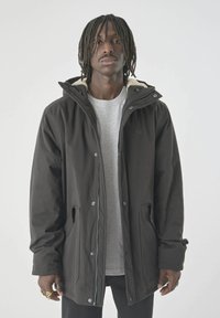 Schwarze isolierte Jacke mit Kapuze, ausgestattet mit Druckknöpfen und einem weichen Innenfutter. Getragen über einem grauen Hemd, vor einem hellen Hintergrund präsentiert.