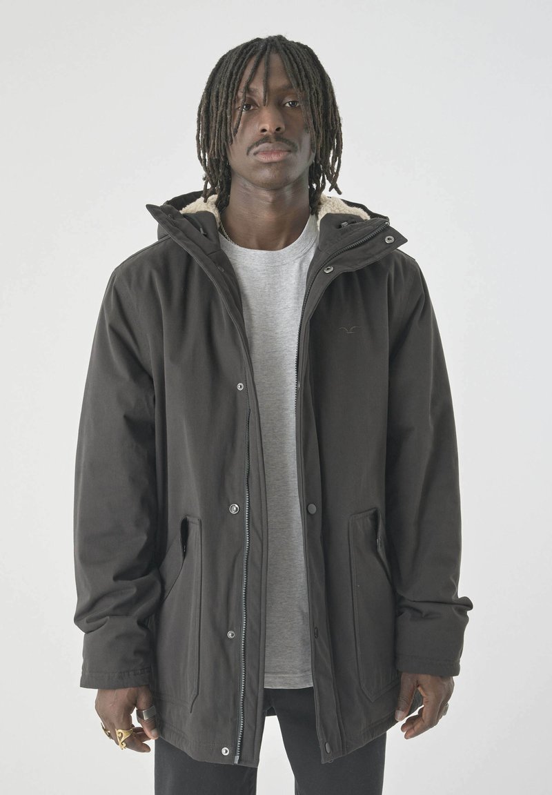Schwarze isolierte Jacke mit Kapuze, ausgestattet mit Druckknöpfen und einem weichen Innenfutter. Getragen über einem grauen Hemd, vor einem hellen Hintergrund präsentiert.