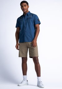 Korte mouw blauwe shirt met knoopsluiting aan de voorkant en twee borstzakken, gecombineerd met tan shorts en witte sneakers, tegen een effen achtergrond.