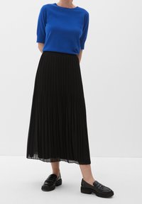 Kvinna som bär en blå t-shirt med kort ärm, svart plisserad midikjol och svarta läderskor med flat klack mot en ren vit bakgrund.