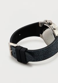 Κοντινό πλάνο ενός ρολογιού χειρός με μαύρο λουράκι από σιλικόνη με υφή και ασημί μεταλλική πόρπη, σε λευκό φόντο.