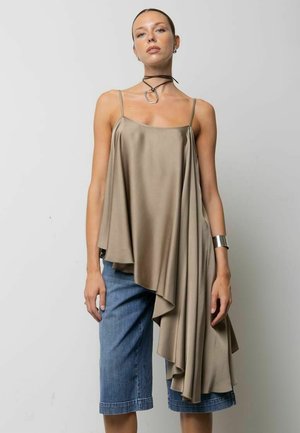 Donna che indossa una camisole asimmetrica color taupe in raso drappeggiato con spalline sottili e pantaloncini di jeans blu al ginocchio su sfondo semplice.