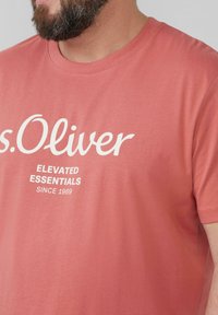 Korallenfarbens Baumwoll-T-Shirt mit Rundhalsausschnitt. Mit weißem Drucktext: "s.Oliver ELEVATED ESSENTIALS SINCE 1969." Schlichtes Design.