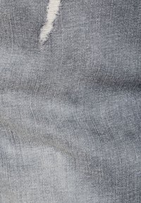 Tejido de mezclilla en azul medio con una textura suave, que presenta un desgarro deshilachado visible. La superficie tiene un sutil patrón de grano.