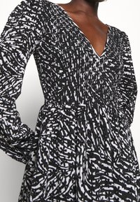 Černobíle vzorované šaty s výstřihem do V, dlouhými rukávy a nabraným pasem. Tkanina má texturované květinové vzory.