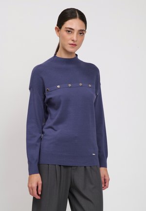 Frau mit dunklen, zurückgebundenen Haaren, die einen langärmeligen violetten Pullover mit Metallknöpfen auf der Brust und lockere graue Hose trägt.
