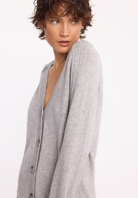Szary kardigan wykonany z miękkiej dzianiny, z dekoltem w serek, z przodu na guziki, długimi rękawami i prążkowanymi akcentami na dolnym brzegu oraz mankietach.