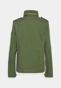 Oliven grøn militærstil jakke med høj krave, guld lynlås og skulderepauleter. Har en indsnævret talje og glat tekstur.