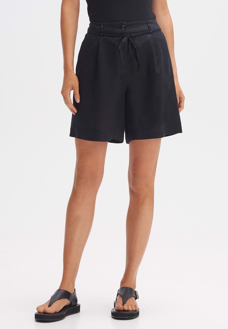 Shorts en lin noir avec une taille à cordon de serrage, des plis à l'avant et des poches latérales. Associés à des sandales noires avec une lanière au niveau des orteils.