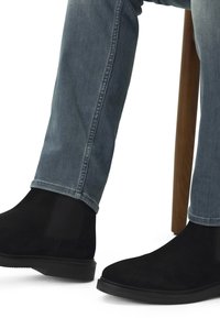Bottines en daim noir avec des panneaux élastiques sur les côtés, associées à un jean en denim bleu clair, assises à côté d'un tabouret en bois.