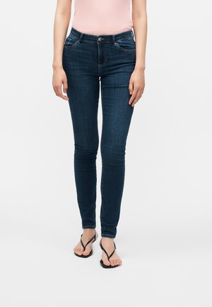 Person iført mørkeblå skinny jeans, en lyserød ærmeløs top og sorte klipklapper, står mod en ensfarvet hvid baggrund.