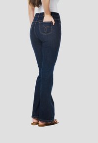 Donkerblauwe uitlopende spijkerbroek met achterzakken, voorzien van een subtiel rond stikselpatroon. Draagt bruine sandalen, gezien van opzij.