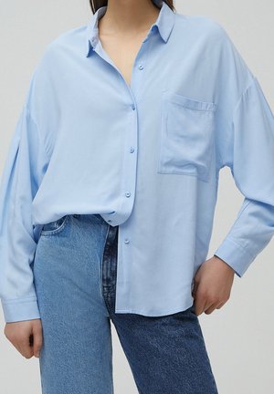 Woman wearing a loose light blue button-up shirt tucked into high-waisted two-tone blue jeans with one hand in pocket.