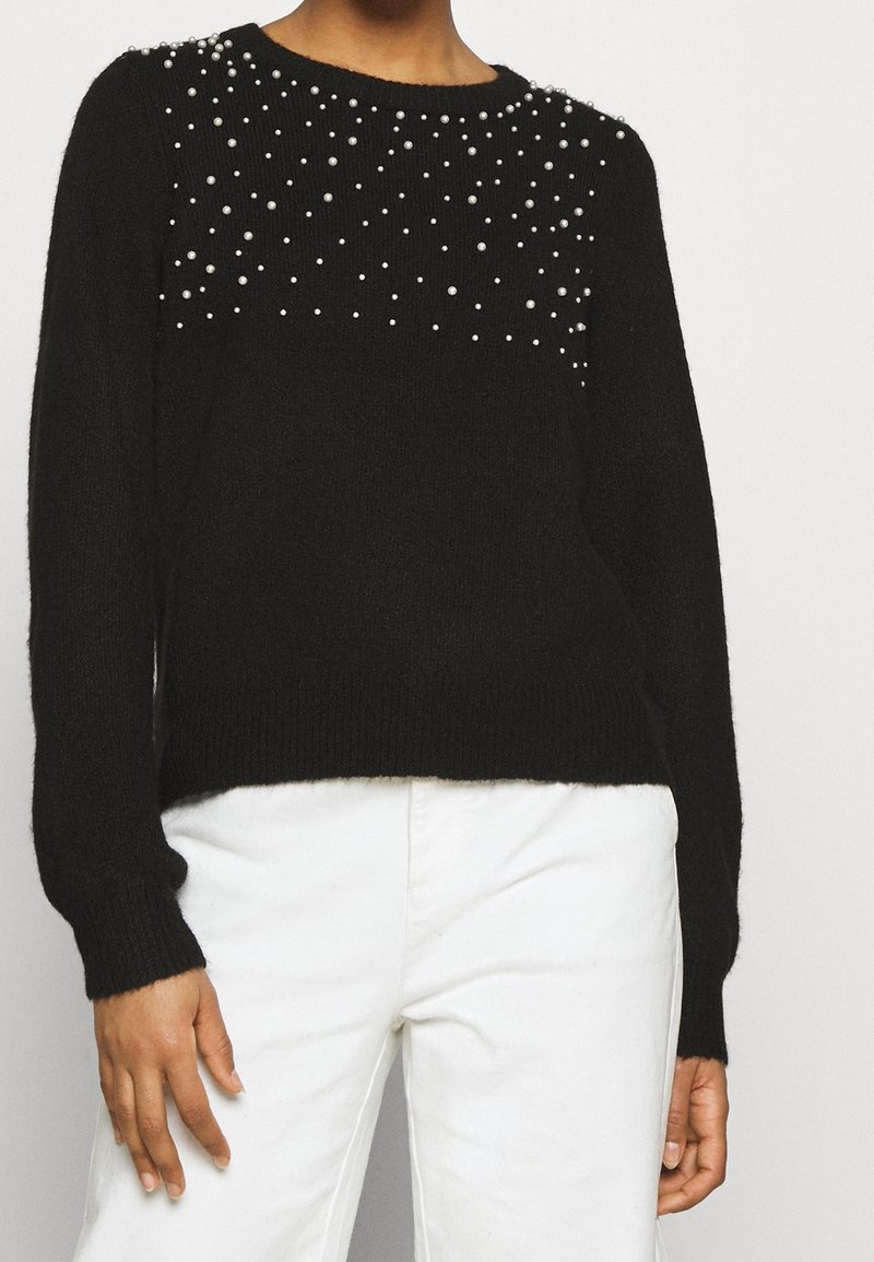 Svart långärmad stickad tröja med spridda pärldekorationer på övre bröstet och axlarna, kombinerad med högmidjade vita byxor.