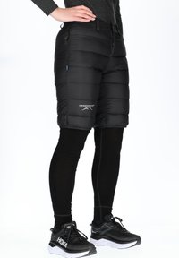 Black insulated shorts with a quilted design, featuring zippered pockets and a logo on the front. Worn over black leggings and sneakers.