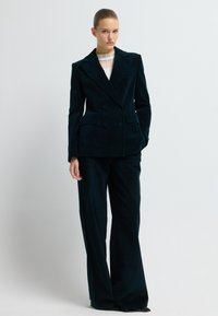 Žena v černém dvouřadovém blejzru a odpovídajících širokých kalhotách, stojící na pozadí jednoduchého světlého pozadí.