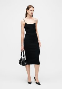 Γυναίκα με μαύρο μίντι φόρεμα με λουράκια από αλυσίδα, κρατώντας μια μαύρη τσάντα με τρουκς και φοράει μαύρες μυτερές ψηλοτάκουνες γόβες.