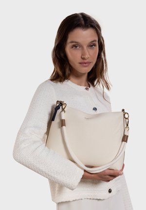 Sac épaule en toile crème avec poignée en corde, ferrures dorées et accents beige. Porté par un mannequin portant un haut blanc texturé.