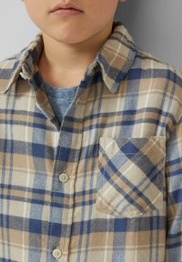 Jonge kind met een beige, blauwe en witte geruite overhemdbroek met een voorzak over een grijze ronde hals ondershirt, tegen een effen achtergrond.