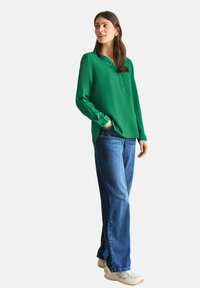 Blusa verde de manga larga con cuello y detalle de botones, combinada con jeans rectos azules y zapatillas beige. Textura de tela suave.