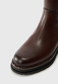 Καφέ δερμάτινο μποτάκι με υφή στην επιφάνεια, ραμμένη σόλα και μαύρη ελαστική σόλα, εμφανίζεται από την εμπρόσθια πλευρά σε λευκό φόντο.