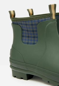 Zielony gumowy botek za kostkę z niebieską tkaniną w kratę wewnątrz, posiada pętle do wkładania, teksturowaną podeszwę i gładkie wykończenie po bokach.