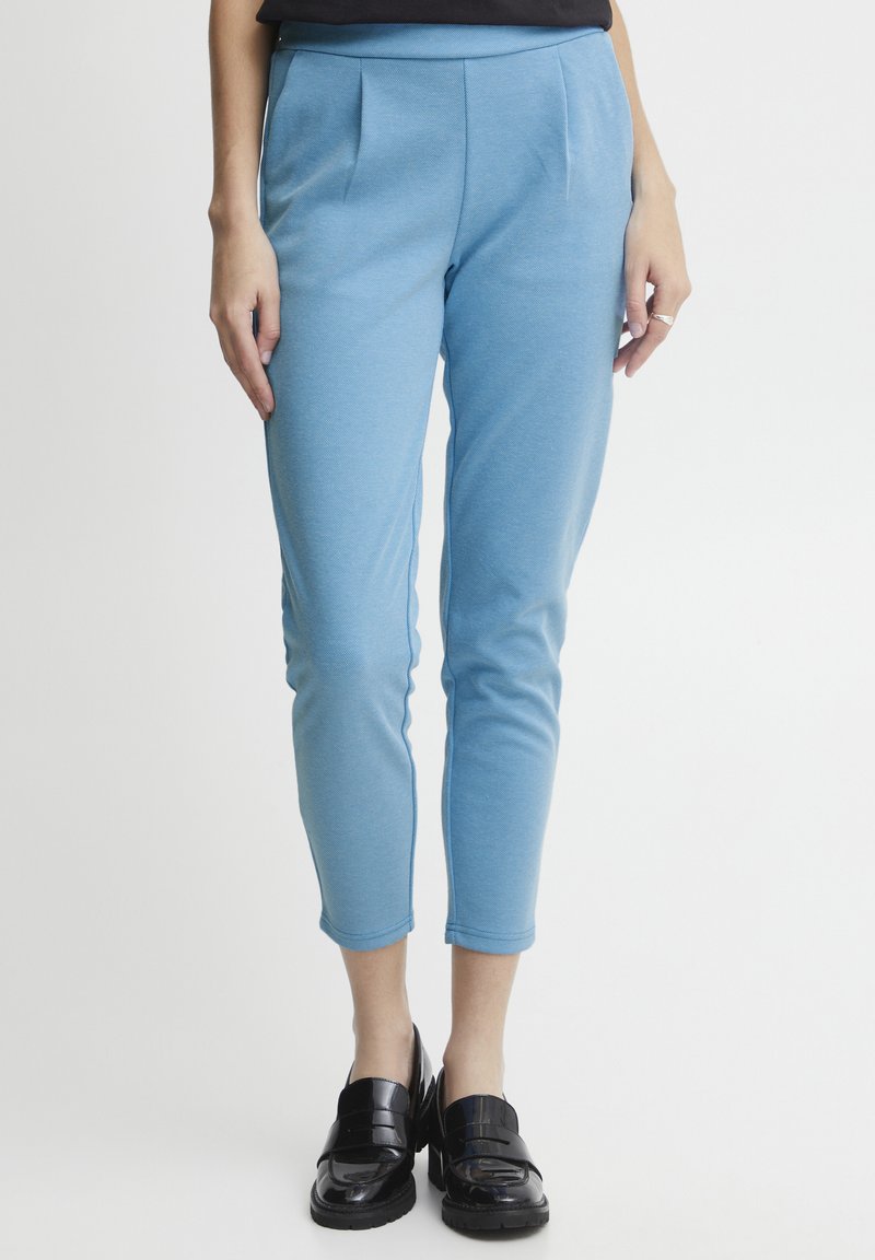 Pantalon ajusté bleu avec des plis à l'avant, fabriqué en un tissu doux et texturé. Longueur courte, associé à des mocassins noirs brillants.