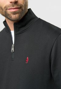 Crna zip-up flis jakna s visokim ovratnikom, koja sadrži mali crveni izvezeni logo na lijevoj strani prsiju i glatku teksturu.