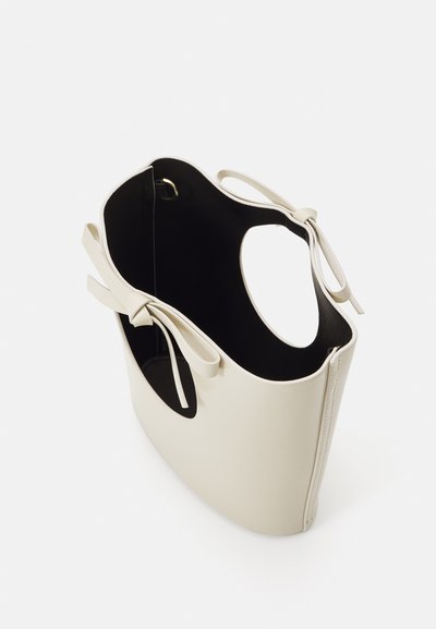 Cream faux leather tote bag with a smooth finish, open top, and two decorative ties on each side. Interior lined in black.