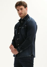 Donkerblauw denim shirt met lange mouwen, knoopsluiting, twee borstzakken en een iets afgeronde zoom. Gladde textuur met subtiele vervaging.