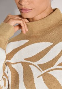 Nahaufnahme einer Person, die einen beigen und weißen gemusterten Rollkragenpulli trägt und sanft den Kragen berührt.