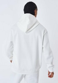 Sudadera blanca hecha de una tela suave, con capucha ajustable, hombros caídos y puños y dobladillo acanalados. Textura lisa sin patrones.