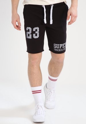Mand iført sorte shorts med hvide snore, trykt "23" og "SUPER TRACK & FIELD," hvide sokker med røde striber og hvide sneakers, der går.