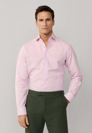 Homme portant une chemise rose clair à boutons et un pantalon vert foncé, debout avec une main dans la poche devant un fond uni.