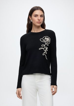 Woman wearing black long-sleeve sweater with white abstract floral embroidery, paired with white pants against a plain white background.