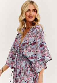 Dlhé šaty vo voľnom štýle v svetlomodrej farbe s paisley vzorom v ružovej, červenej a čiernej farbe. Obsahujú široké rukávy a elastický pás.