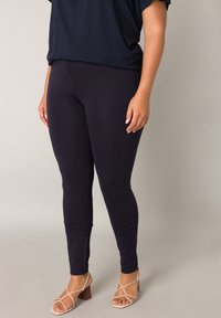 Vrouw die zwarte nauwsluitende broek en beige sandalen met blokhak en bandjes draagt, tegen een effen lichte achtergrond.