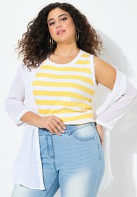 Randig gul och vit linne under en vit, genomskinlig skjorta med knappar. Bärs med ljusblå denimjeans. Ljus från tidig eftermiddag.